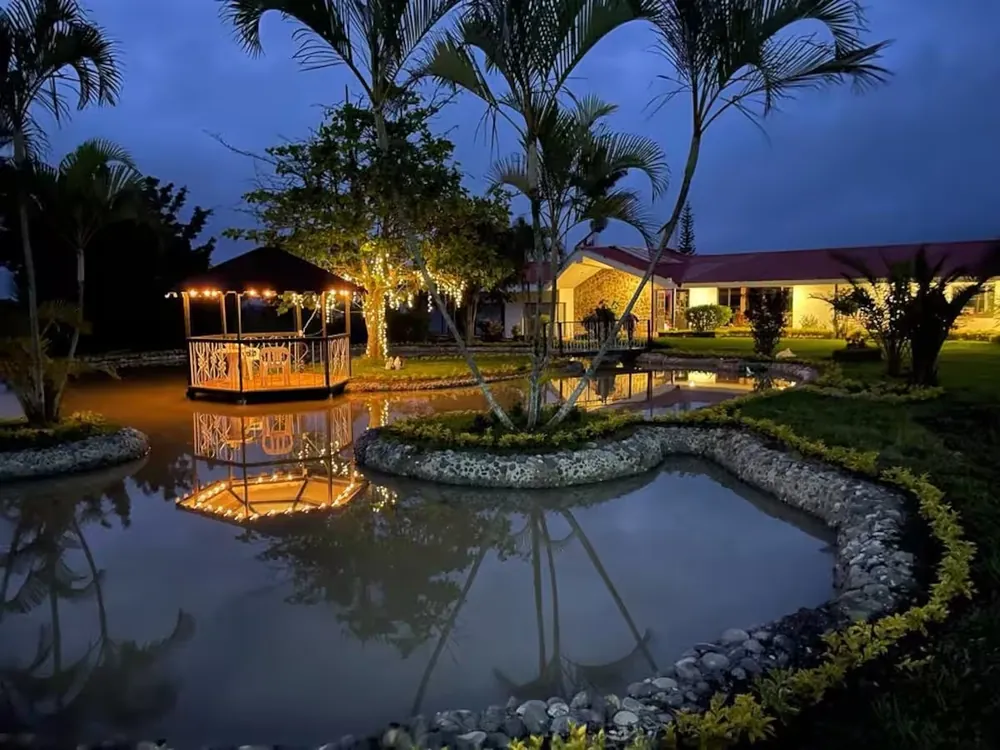 La casa campestre de noche: un kiosco con luces junto a un estanque, palmeras y la casa al fondo